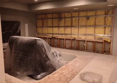 An indoor room under renovation, with exposed wall studs and insulation on one wall, and a large object covered in plastic in the foreground. The floor appears to be concrete or subfloor.