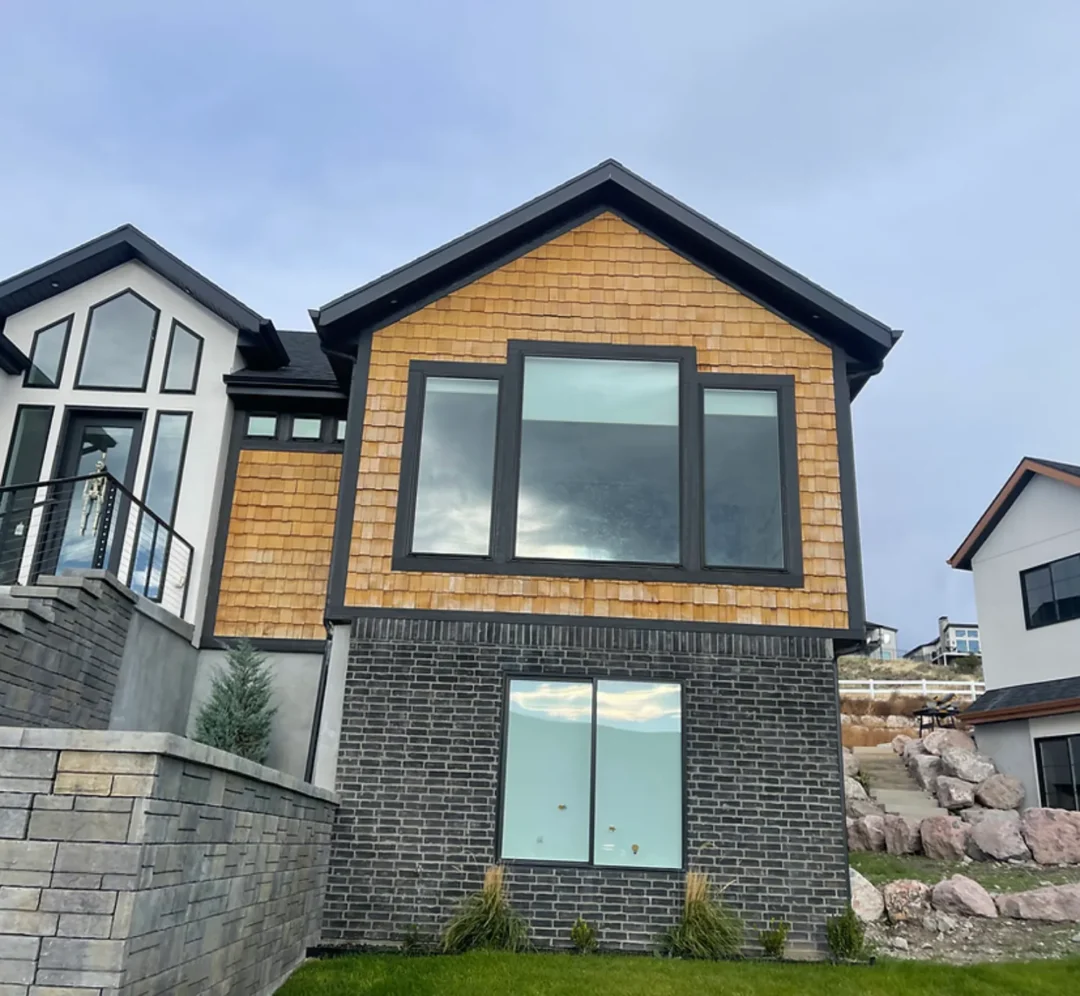 The front exterior of a modern multi-story house with dark grey brick on the first floor and horizontal wood paneling on the second, featuring large black-framed windows and a distinct roofline.