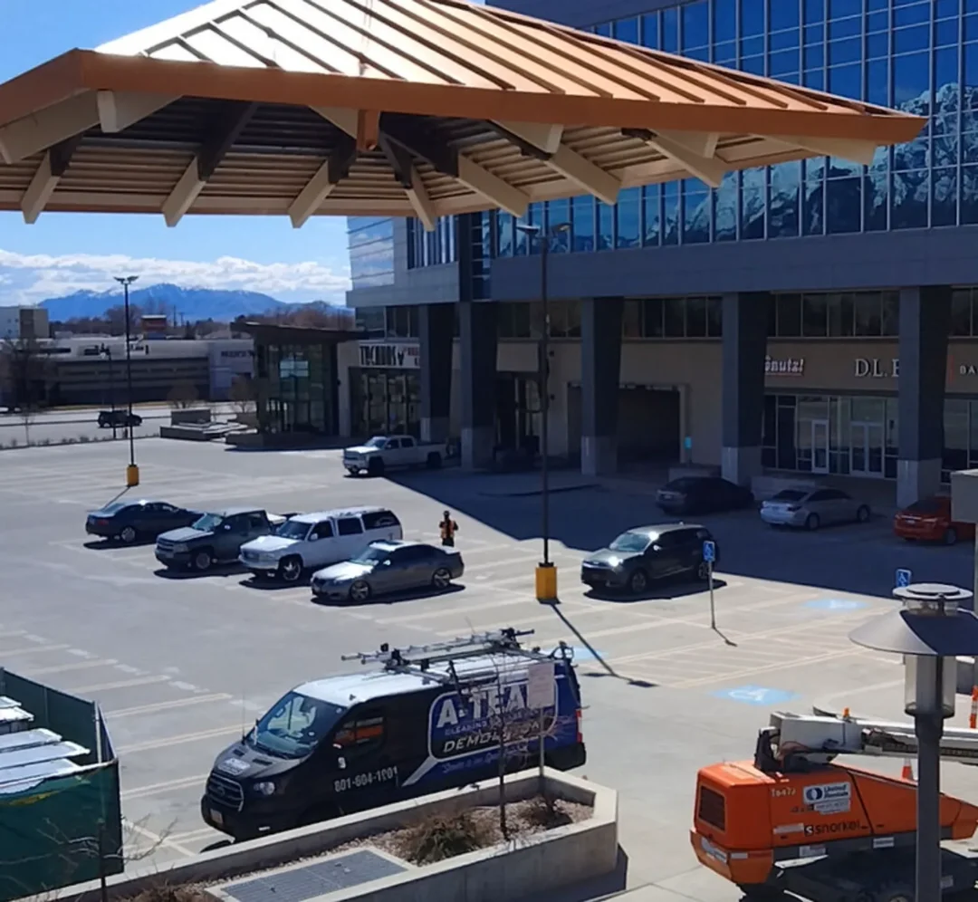 A wide shot of a parking lot in front of a modern glass building with a distinct wooden awning, showing several parked cars, a black van with a "A-TEAM DEMO" logo, and a small orange lift vehicle. Mountains are visible in the background.